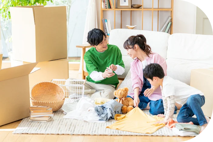 引っ越しの荷造りをしている男女と男の子。部屋には段ボール箱が積まれ、床には衣類やおもちゃが広げられている。3人は笑顔で会話している。