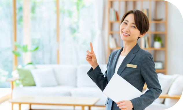 スーツを着用し、白い書類を手に持って指で指し示しながら微笑んでいる女性。背景にはリビングが広がっている。