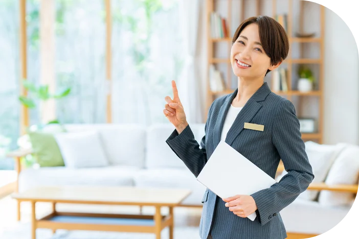 スーツを着用し、白い書類を手に持って指で指し示しながら微笑んでいる女性。背景にはリビングが広がっている。